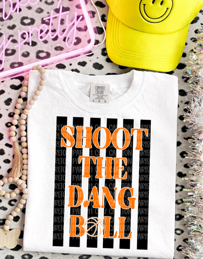White t-shirt with 'SHOOT THE DANG BALL' text on a patterned surface with accessories.