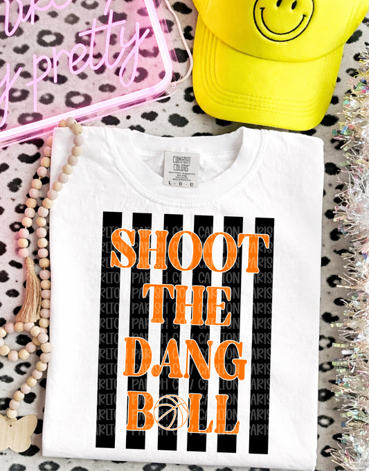 White t-shirt with 'SHOOT THE DANG BALL' text on a patterned surface with accessories.