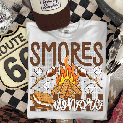 White t-shirt with 'S'mores Whore
' design featuring a campfire and marshmallows, placed on a patterned surface.